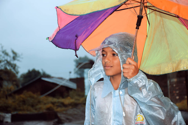 ein indischer Junge, der wegsieht, Regenbogen im nassen Wasser trägt, mit buntem Regenschirm, Blick auf das Dorf - Foto, Bild