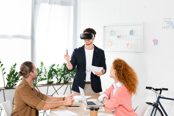 smiling businessman with virtual reality headset holding pen and papers and friends looking at him  - Photo, Image