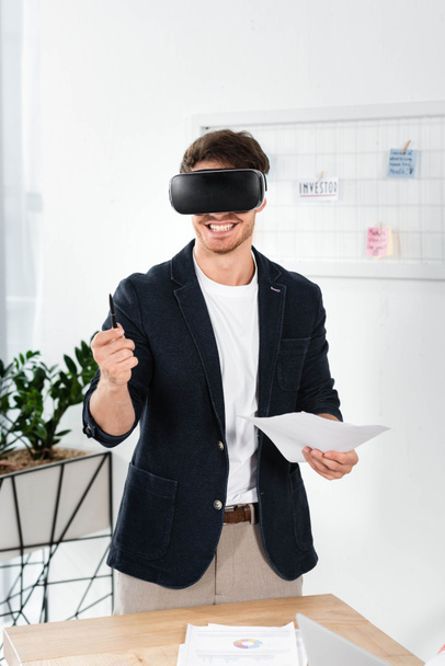 smiling businessman in shirt with virtual reality headset holding pen and papers - Photo, Image