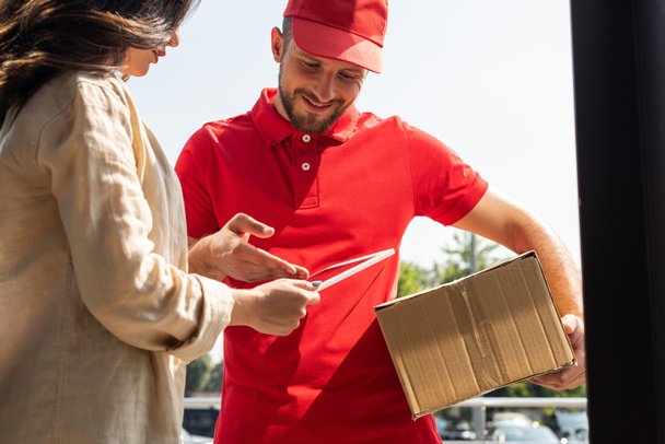 cheerful delivery man gesturing while looking at digital tablet near woman  - Photo, Image