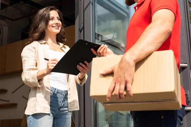 cropped view of delivery man holding box near attractive girl with clipboard  - Foto, afbeelding