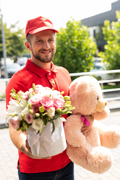 selective focus of cheerful delivery man holding teddy bear and flowers  - Photo, Image
