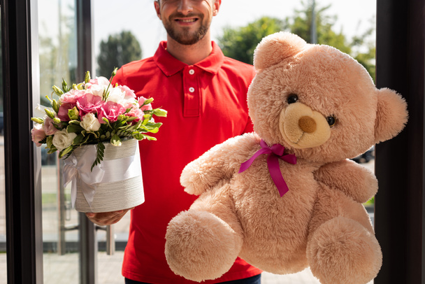 cropped view of positive delivery man holding teddy bear and flowers  - Φωτογραφία, εικόνα