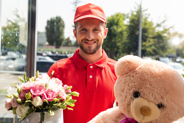 Lächelnder Lieferant mit Mütze und Teddybär und Blumen  - Foto, Bild
