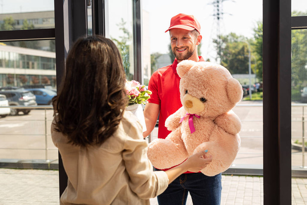 selective focus of happy delivery man in cap holding teddy bear and flowers near woman  - Photo, Image
