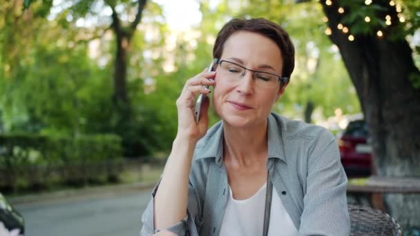 Mature woman in glasses talking on mobile phone in open air cafe outdoors - Záběry, video