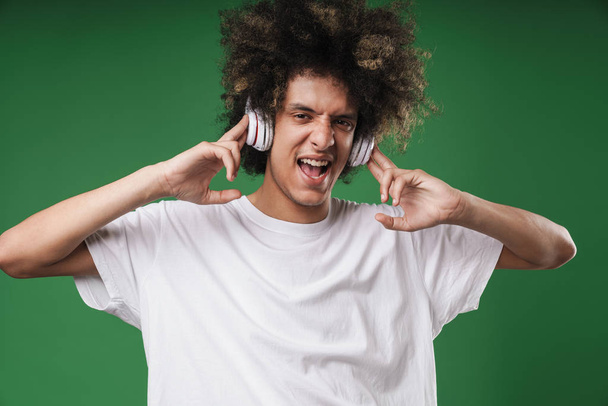 Curly guy posing isolated over green wall background listening music with headphones. - Foto, Bild