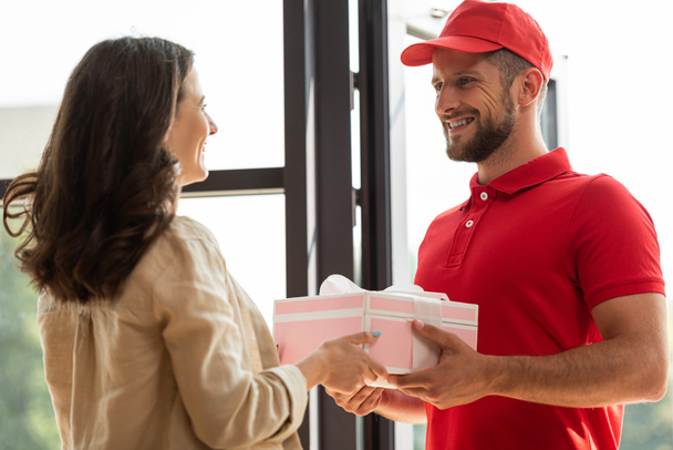 handsome delivery man in cap giving pink present to happy woman  - Fotoğraf, Görsel