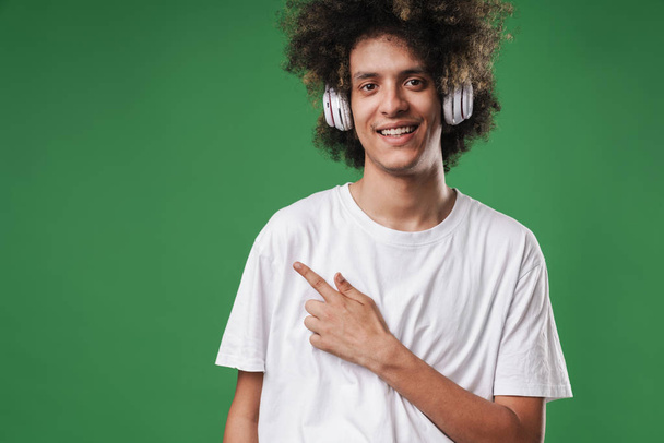 Smiling curly guy posing isolated over green wall background listening music with headphones pointing to copyspace. - Fotoğraf, Görsel