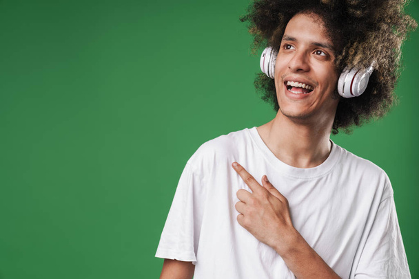 Smiling curly guy posing isolated over green wall background listening music with headphones pointing to copyspace. - Foto, imagen