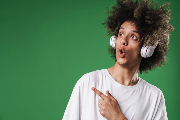 Chico rizado sorprendido posando aislado sobre fondo de pared verde escuchando música con auriculares apuntando al espacio de copia
. - Foto, imagen