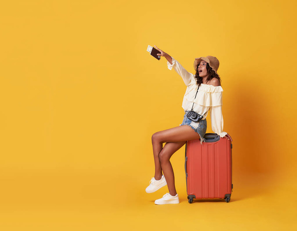 Cheerful young african woman dressed in summer clothes sitting on a suitcase and pointing finger at copy space isolated over yellow background. - Photo, Image