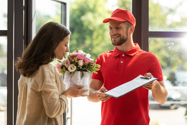 bearded and happy delivery man holding clipboard near beautiful woman smelling flowers  - Photo, Image