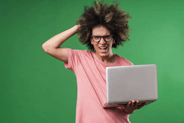 Young angry screaming curly guy posing isolated over green wall background using laptop computer. - Photo, Image