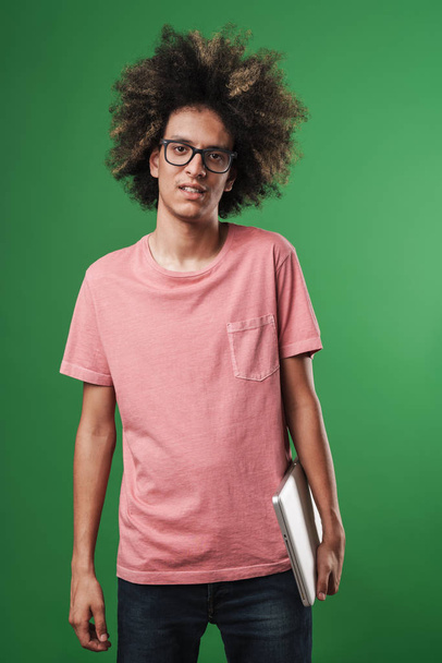 Young curly guy posing isolated over green wall background holding laptop computer. - Photo, Image