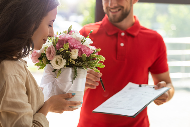 selective focus of woman smelling flowers near delivery man holding clipboard - Foto, immagini