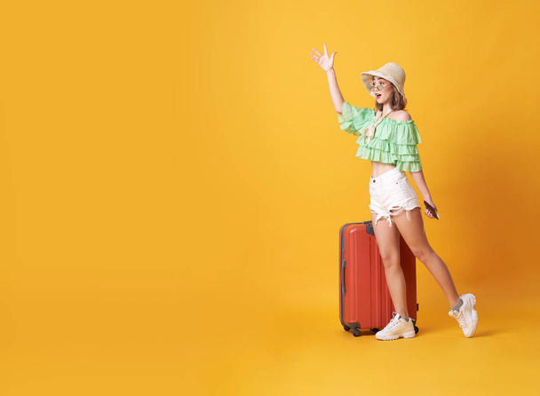 Веселая молодая женщина, одетая в летнюю одежду, стоит с чемоданом и здоровается на копировальном пространстве, изолированном на желтом фоне
. - Фото, изображение