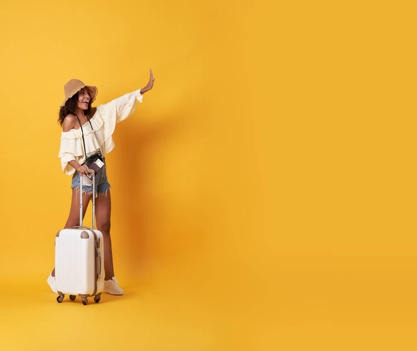 Vrolijke jonge Afrikaanse vrouw gekleed in zomer kleren staan met een koffer en zeggen hallo op kopie ruimte geïsoleerd over gele achtergrond. - Foto, afbeelding