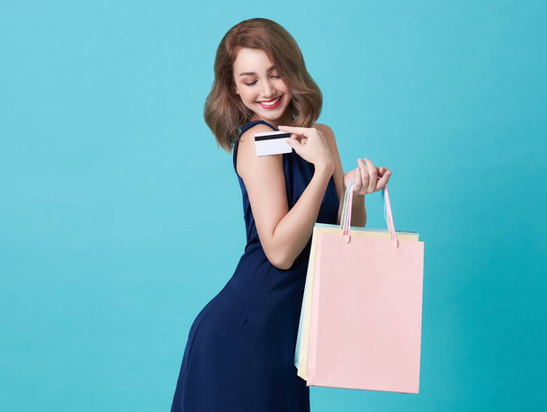 Πορτραίτο μιας ευτυχης νεαρής γυναίκας που δείχνει πιστωτική κάρτα και τσάντα για ψώνια απομονωμένα πάνω από μπλε φόντο. - Φωτογραφία, εικόνα