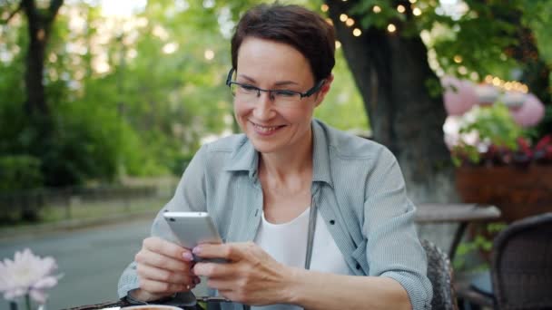 Smiling lady using smartphone touching screen smiling in street cafe outside - Séquence, vidéo