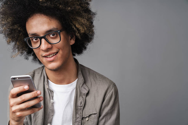 Photo closeup of young pleased man with afro hairstyle smiling at camera and using cellphone - 写真・画像