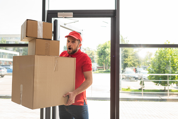 shocked and bearded man holding carton boxes while looking at camera  - Photo, Image