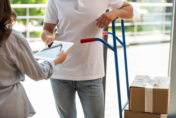 cropped view of delivery man standing near delivery cart with boxes and woman with digital tablet  - Foto, Bild