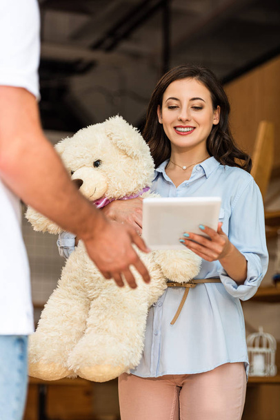 vista de ángulo bajo de entrega hombre gesto cerca de mujer feliz sosteniendo tableta digital y oso de peluche
  - Foto, Imagen