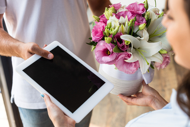 女性に空白の画面で花とデジタルタブレットを与える配達人のトリミングビュー  - 写真・画像