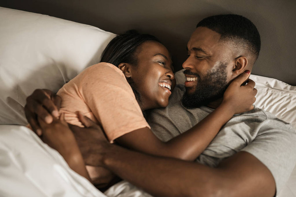 Affectionate young African American couple hugging and laughing together while lying in bed in the morning - Фото, зображення