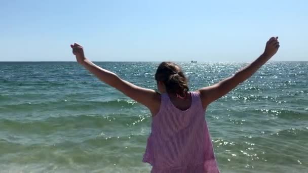 Vista lateral de cerca de una joven mujer caucásica de pie en una playa con sus brazos en el aire, mirando al mar
 - Metraje, vídeo