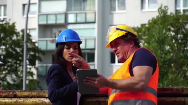 Male and female workers on construction site wearing safety vest, helmet and goggles checking the work plan on a digital tablet computer - Footage, Video
