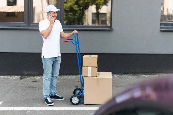 selective focus of happy bearded delivery man talking on smartphone while holding delivery cart  - Photo, Image