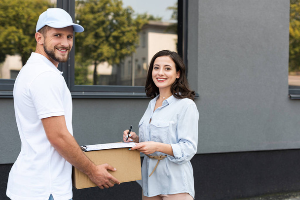 smiling delivery man holding box near woman signing paper on clipboard outside  - Foto, imagen