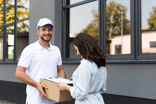 happy bearded delivery man holding box near woman signing paper on clipboard outside  - Foto, Imagen