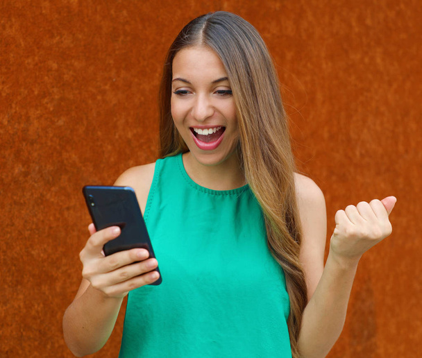 Emocionada joven mujer de negocios que recibe buenas noticias en el teléfono inteligente celebrando con el puño en alto al aire libre
. - Foto, imagen