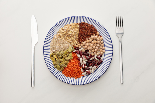 Gestreifter Teller mit rohen Linsen, Kichererbsen, Quinoa, Haferflocken, Bohnen und Kürbiskernen in der Nähe von Besteck auf Marmoroberfläche - Foto, Bild