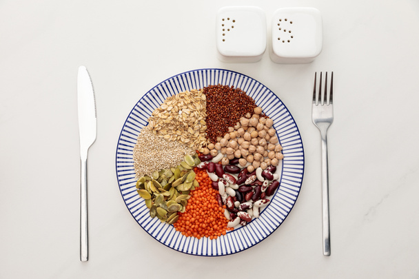 top view of plate with raw cereal, diverse beans and pumpkin seeds near cutlery and salt and pepper pots on marble surface - Photo, Image