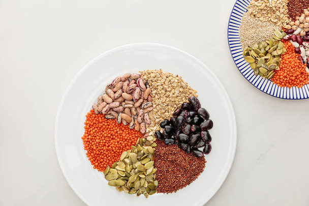 Ansicht von weißen und gestreiften Tellern mit rohen Linsen, Quinoa, Haferflocken, Bohnen und Kürbiskernen auf Marmoroberfläche - Foto, Bild