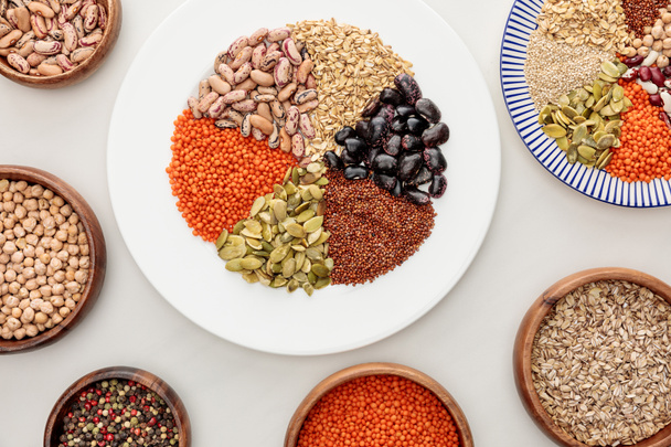 top view of white and striped plates and wooden bowls with raw lentil, quinoa, oatmeal, beans, peppercorns and pumpkin seeds on marble surface - Photo, Image