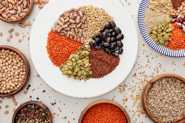 top view of plates and bowls with raw lentil, quinoa, oatmeal, beans, peppercorns and pumpkin seeds on marble surface with scattered grains - Foto, afbeelding