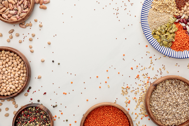 bovenaanzicht van plaat en kommen met rauwe linzen, quinoa, havermout, bonen, peperkorrels en pompoenpitten op marmer oppervlak met verspreide korrels en kopieer ruimte - Foto, afbeelding
