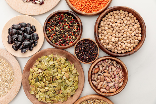 top view of wooden bowls with diverse beans, quinoa, red lentil, peppercorns, pumpkin seeds and chickpea on white marble surface - Foto, immagini