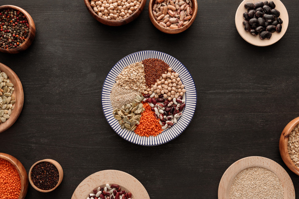 top view of striped plate with various raw legumes and cereals near bowls on dark wooden surface - Foto, imagen