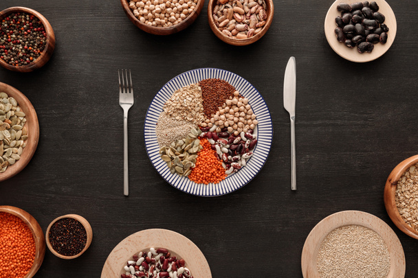 top view of knife and fork near striped plate with various raw legumes and cereals and bowls on dark wooden surface - Fotó, kép