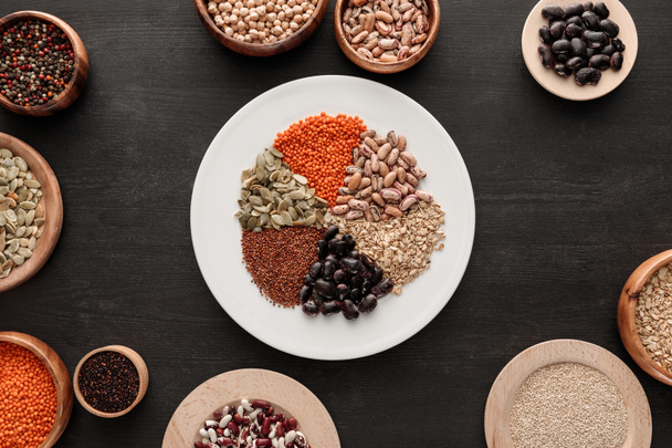 top view of white plate with various raw legumes and cereals near bowls on dark wooden surface - Fotografie, Obrázek