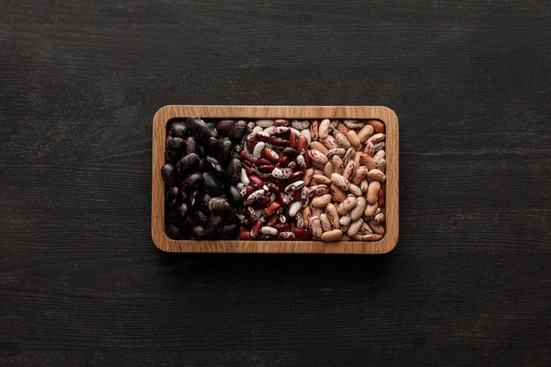 コピースペースを持つ暗い木製の表面に生の盛り合わせ豆と長方形の茶色のボウルのトップビュー - 写真・画像