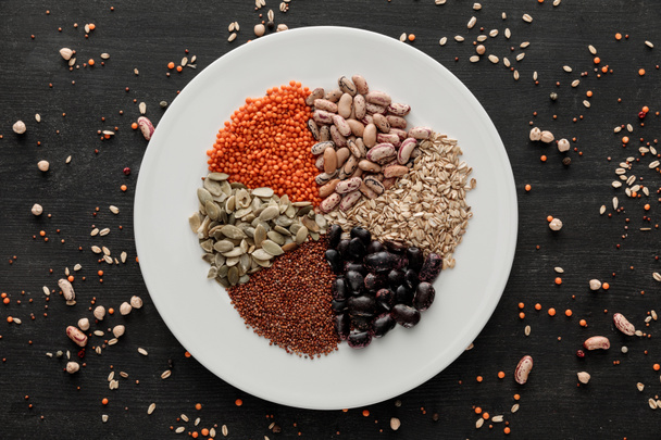 top view of white ceramic plate with raw assorted beans, cereals and seeds on dark wooden surface with scattered grains - Photo, Image