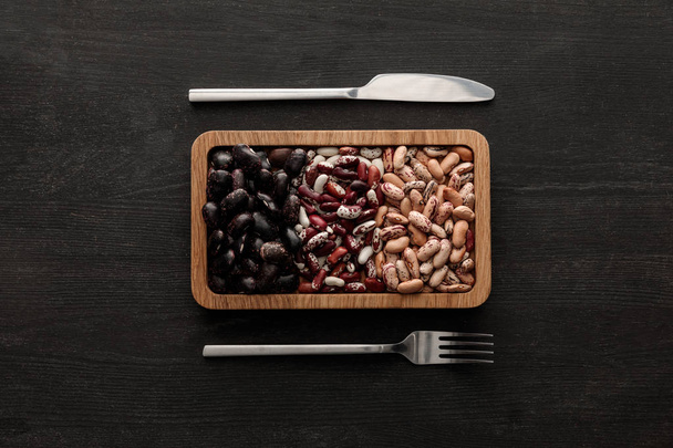 top view of fork and knife near rectangular brown bowl with raw assorted beans on dark wooden surface - Foto, imagen