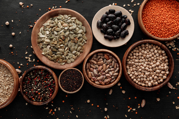 top view of wooden bowls and plates with raw assorted beans, cereals and seeds on dark surface with scattered grains - Foto, imagen